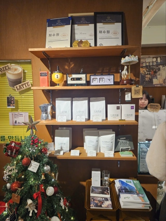 칠복상회로스터스 원두 및 굿즈 진열대: 골드 상장과 원두 패키지, 크리스마스 트리가 있는 모습