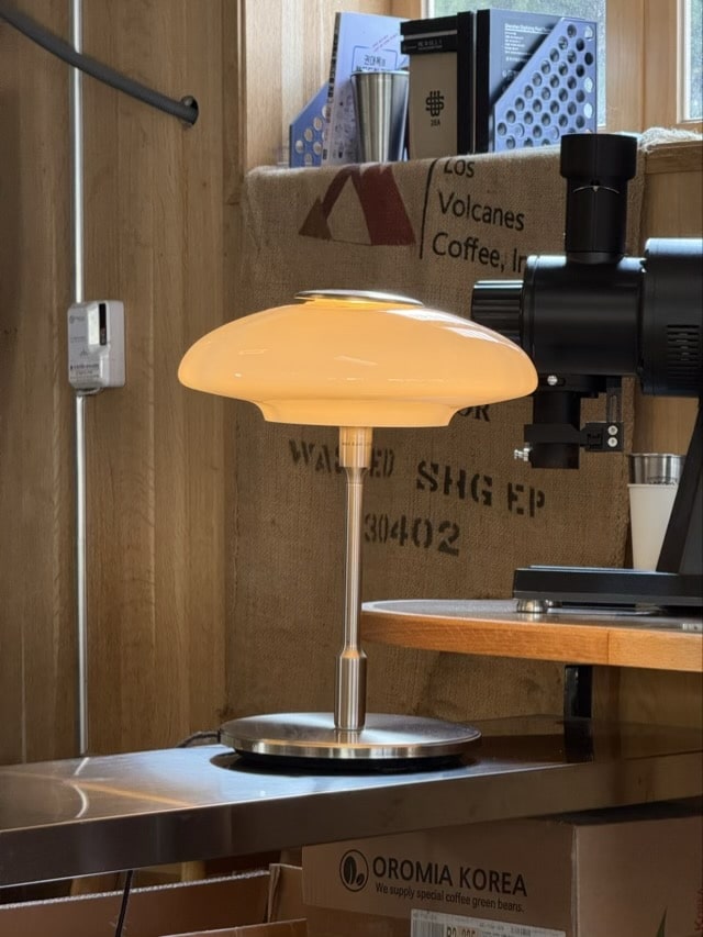 커피 그라인더 옆 스틸 테이블 위에 놓인 모던하면서도 클래식한 디자인의 갓 조명.